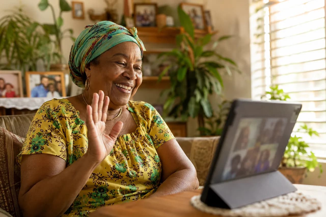 A Jamaican grandmother waving warmly at a tablet on a video call with family overseas
